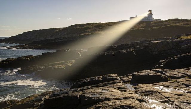 Lighthouse casting golden beam over rocky shoreline and tidal pools at golden hour beacon