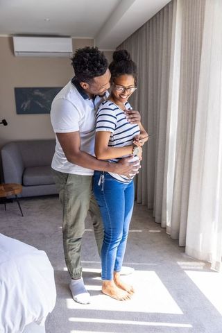 African american couple embracing by window, expecting baby, sunlit bedroom moment
