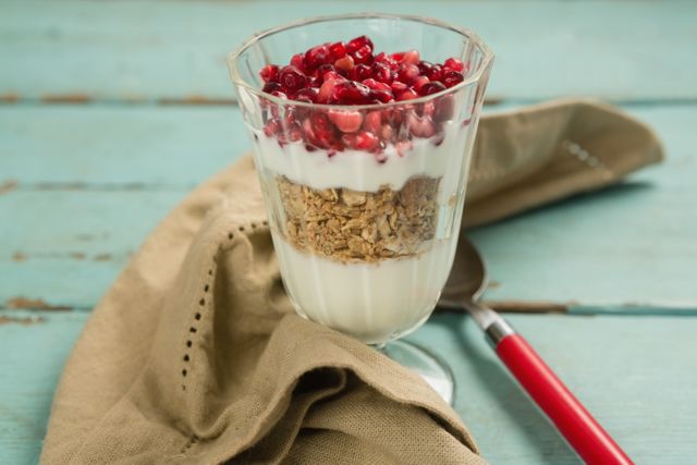Layered yogurt parfait with granola and pomegranate seeds in a glass cup on a rustic wooden table. Ideal for promoting healthy eating, breakfast recipes, or nutritious snacks. Perfect for food blogs, recipe websites, and health magazines.
