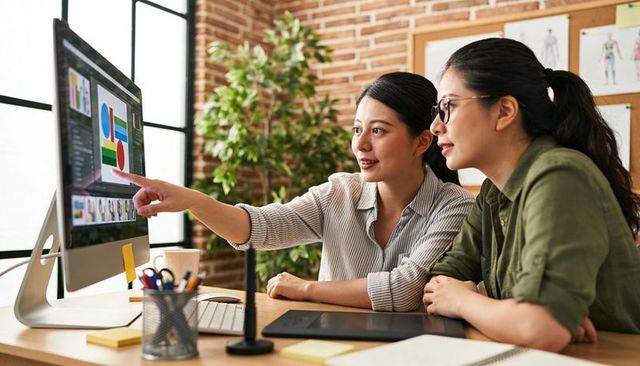 Asian women collaborating on graphic design, reviewing charts on desktop monitor in studio