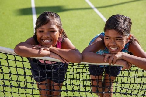 Smiling girls leaning over tennis net in sunlit court