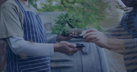 Exchanging bonsai with contactless mobile payment in backyard vendor wearing striped apron