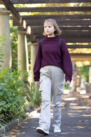 Young woman walking on garden pathway under pergola wearing burgundy sweater and cargo pants