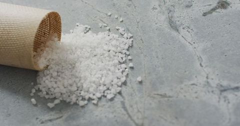 Coarse Sea Salt Spilling from Woven Cup on Stone Surface