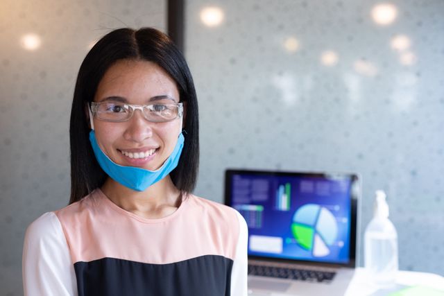 Asian woman wearing face mask and protective glasses in an office environment during the COVID-19 pandemic. She is smiling and standing in front of a laptop displaying charts and graphs, with a bottle of hand sanitizer nearby. Useful for topics related to workplace safety, health measures, pandemic protocols, and professional settings during COVID-19.