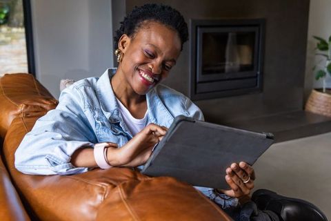 African american woman relaxing on leather sofa using tablet in modern living room with fireplace