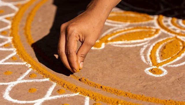 Hand sprinkling turmeric powder drawing intricate rangoli petal on sunlit clay courtyard