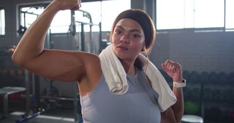 Mid-adult woman flexing biceps in sunlit industrial gym wearing towel, headband, smartwatch