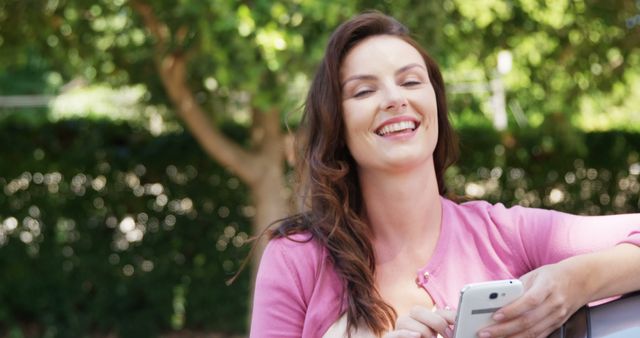 A joyful woman standing outside in a park, holding a smartphone and smiling. She is dressed in a pink cardigan against a backdrop of lush greenery, embodying relaxation and the joy of outdoor leisure time. Perfect for use in advertisements for mobile technology, park activities, relaxation apps, or lifestyle blogs. Illustrates carefree enjoyment and digital connectivity in nature.