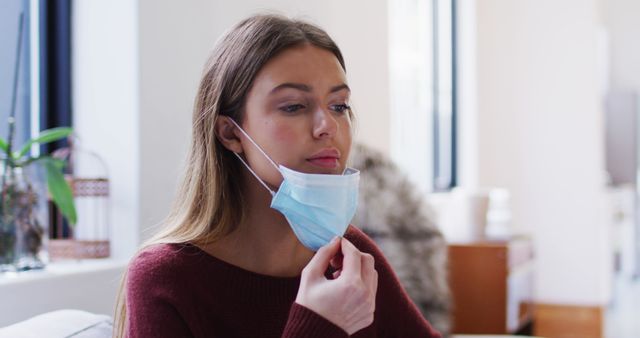 Young woman fixing her face mask while sitting in a room with a soft and bright ambiance. Use this image for health awareness, pandemic-related content, or articles emphasizing the importance of wearing masks to prevent disease spread.