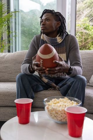Pensive african american man sitting on couch holding football while snacking on popcorn