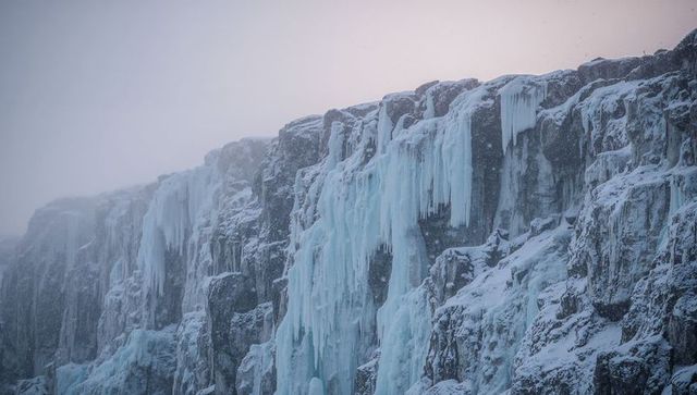 Frozen cliff draping cascading turquoise ice and icicles, snow falling through mist