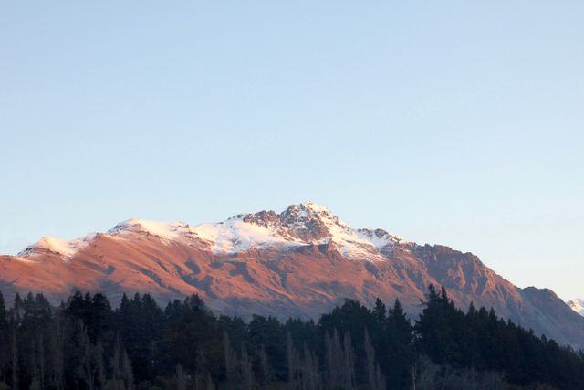 This beautiful image captures a snow-capped mountain peak being illuminated by the first light of dawn, with a dense forest in the foreground. This scene is perfect for use in promotional materials for travel and adventure, nature publications, or websites focused on outdoor activities. It evokes tranquility, awe, and the majestic beauty of natural landscapes.