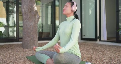Asian woman meditating on patio with pink headphones and yoga mat for mindfulness, wellness