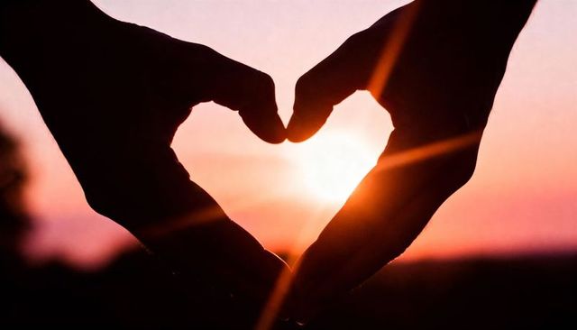 Silhouetted hands forming heart around sun at sunset with warm lens flare and glow