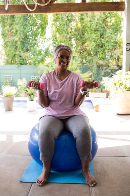 Senior woman sitting on a blue fitness ball, holding pink dumbbells, and exercising in a garden. She is wearing a pink shirt and gray leggings, smiling and looking healthy. Ideal for use in articles or advertisements promoting senior fitness, healthy lifestyles, home workouts, and wellness programs.