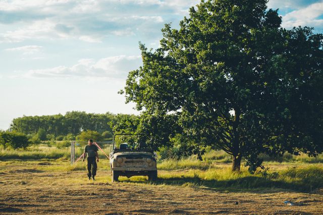 An individual is walking towards an abandoned car beneath a large tree in an open countryside field. Green grass and clear skies create a serene scene ideal for concepts related to solitude, exploration, or rural life. Useful for illustrating themes of adventure, nature retreats, or rural living.