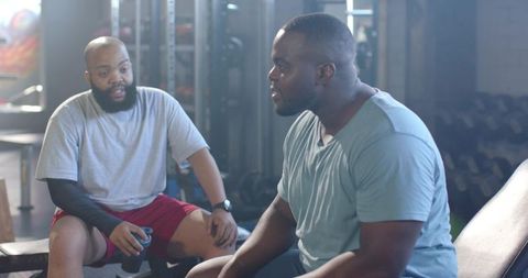 Two african men talking while resting on gym bench after strength workout with shaker bottle