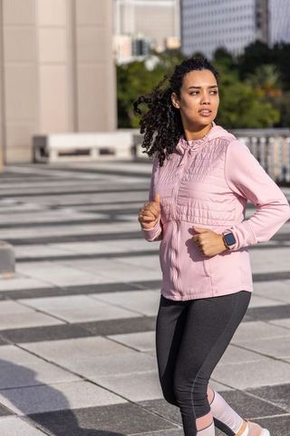Sunlit urban runner jogging on rooftop terrace wearing pink jacket and smartwatch