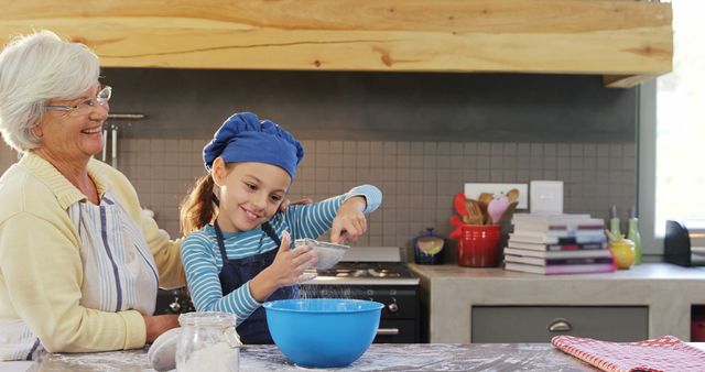 Grandmother and granddaughter enjoying baking together in a modern kitchen. Granddaughter wearing a blue chef hat and apron, sieving flour into a bowl, while grandmother smiles. Ideal for content on family activities, cooking with kids, bonding moments, and healthy family lifestyles.