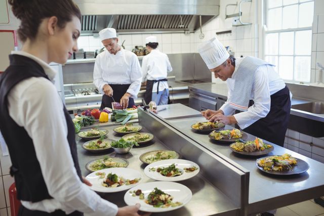 Waitress collecting dishes from order station while chefs are busy preparing and plating food in a professional restaurant kitchen. Ideal for use in content related to culinary arts, restaurant operations, hospitality industry, teamwork in kitchens, and food service training.