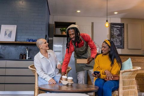 African american barista serving coffee to friends in cozy cafe with wicker chairs