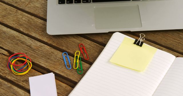 Open notebook with a yellow sticky note and a binder clip, colorful paper clips and rubber bands, along with a laptop on a wooden table. Ideal for illustrating concepts of workspace organization, office work, studying, remote work, and productivity.