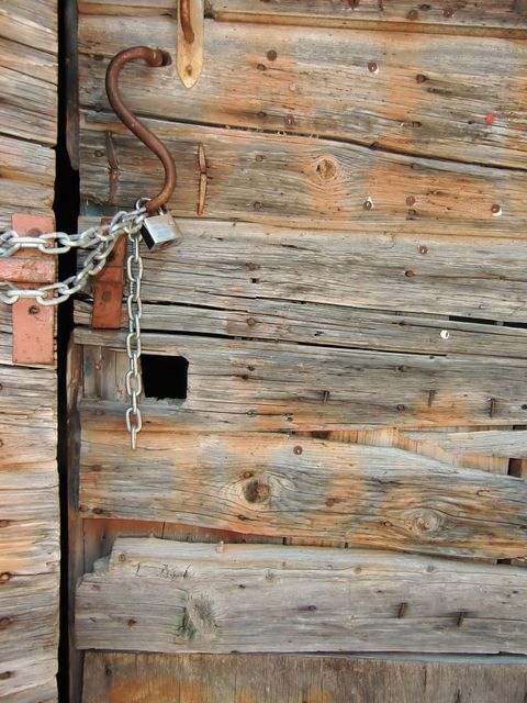 Close-up showing an old wooden door secured with a metal chain and padlock. Details highlight textures of weathered wood, rusted latch, and signs of age. Ideal for themes around security, history, rustic settings, and vintage decor.
