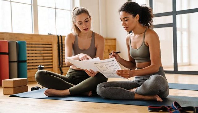 Two women reviewing workout plan on yoga mats in bright studio practicing fitness planning