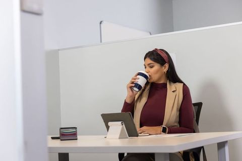 Asian businesswoman drinking coffee and checking tablet in minimalist office cubicle
