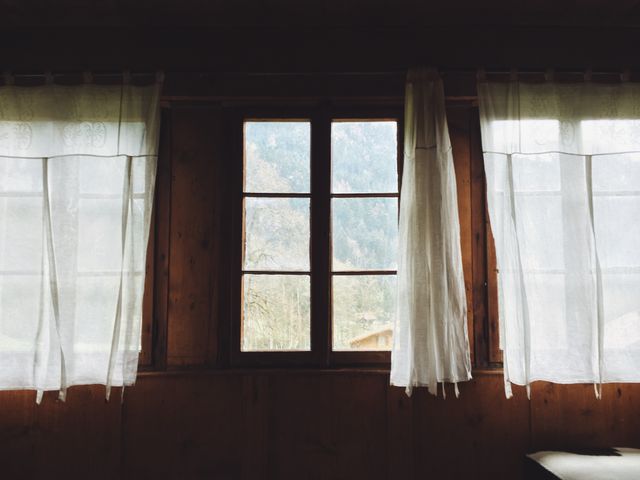 This image shows a rustic wooden window with white curtains, bringing in natural light to the cozy interior of a cabin. Ideal for topics related to home decor, country living, outdoor retreats, nature, and vintage aesthetics.