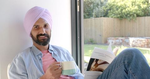 Man in Turban Drinking Coffee and Reading Book by Window