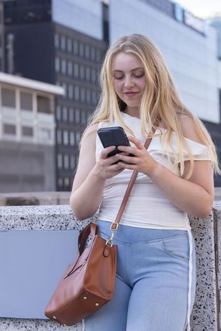 Young woman texting on smartphone on city terrace leaning on railing with crossbody bag
