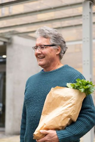 Smiling senior man carrying paper bag of fresh greens walking urban market in teal sweater