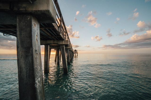 Wooden pier extending over a calm ocean during sunset with scattered clouds in the sky. Ideal for depicting serene beach scenes, travel destinations, relaxation, and coastal landscapes.