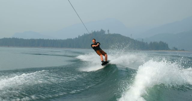 Man wakeboarding on a lake with scenic views of forested mountains in the background. This can be used for promoting outdoor recreational activities, adventure tourism, or summer sports. Perfect for brochures, websites, or advertisements focused on nature and extreme sports.