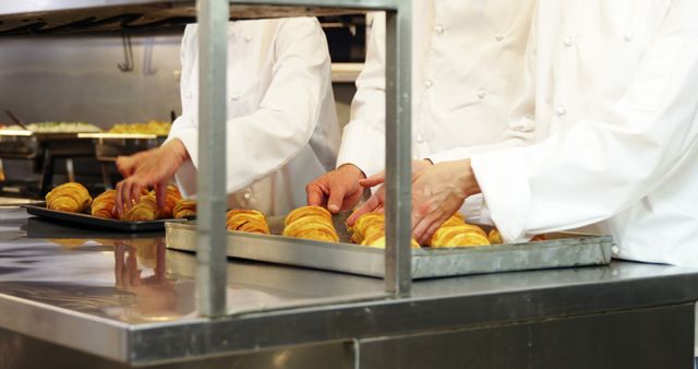 Chefs in white uniforms arranging freshly baked croissants on trays in a professional kitchen. Shows teamwork and precision in culinary arts. Ideal for content related to food industry, professional cooking, culinary schools, and bakery promotions.