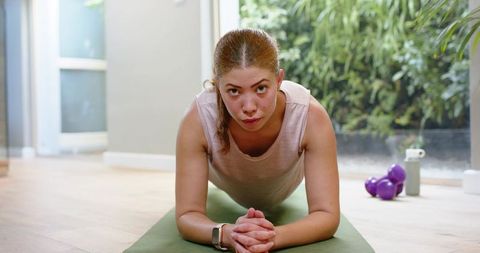 Young woman doing forearm plank on green mat at home workout with purple dumbbells