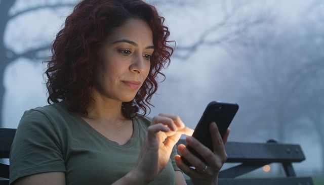 Tapping smartphone while sitting on park bench in foggy evening with warm screen glow