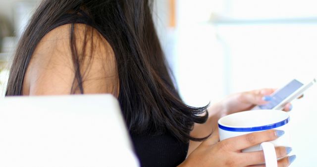 This image captures a woman with long hair holding a coffee mug in one hand and a smartphone in the other. It is perfect for promoting a relaxed lifestyle, morning routines, or technology use at home. Ideal for blogs, articles, or marketing materials concerning daily routines, coffee lovers, or phone applications.