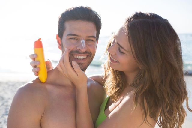 Happy woman applying sunscream on smiling man face at beach on sunny day