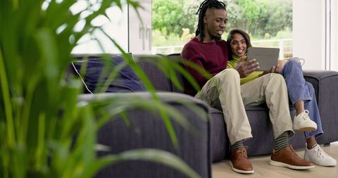 African American man and Indian woman relaxing on sofa sharing tablet in living room