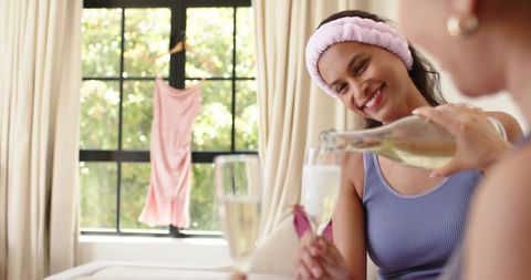 Multicultural women toasting with champagne, relaxing on bed in sunlit bedroom