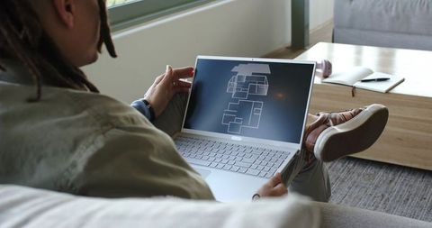 Male with dreadlocks designing modern home floor plan on laptop while lounging on couch