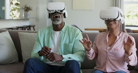 Senior couple laughing and exploring virtual reality on sofa with white VR headsets