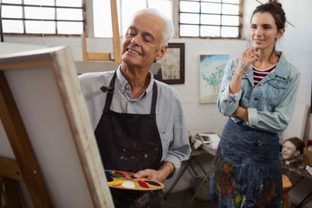 Senior man painting on canvas while a woman observes in an art class. Ideal for use in articles or advertisements about art education, creative workshops, senior activities, and intergenerational learning. Perfect for illustrating the joy of artistic expression and the mentor-student relationship in a creative setting.