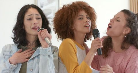 Diverse female friends singing together on couch with microphones home karaoke party