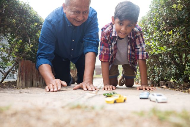 Grandfather and grandson enjoying quality time together by playing with toy cars on a pavement in a yard. Ideal for use in family-oriented advertisements, articles on family bonding, childhood memories, and outdoor activities. Perfect for illustrating multigenerational relationships and leisure activities.