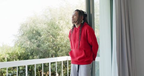 African american man standing on balcony in red hoodie looking out over lush greenery