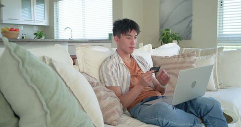 Young asian man lounging on sofa holding credit card and using laptop for online shopping